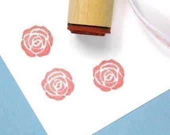 May include: A wooden handled stamp with a red rubber rose design. Three rose impressions in light red are on white paper. A white and pink ribbon is in the background. The stamp is partially visible.