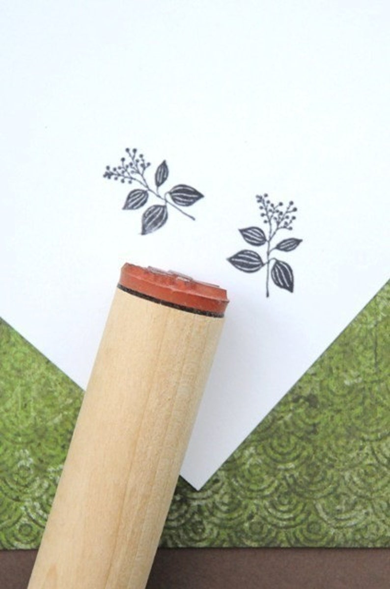 May include: A wooden stamp with a botanical design, featuring a sprig with leaves and small berries, is shown next to two stamped impressions of the same design. The stamp has a red rubber base and a light-colored wooden handle. The background is white and green.