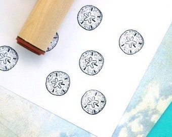 May include: A wooden stamp with a brown rubber base is positioned near several stamped images of sand dollars. The sand dollar images are black and white, with a detailed design. The background is white with hints of blue and teal.