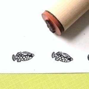 May include: A wooden handled stamp with a fish design. The stamp has a detailed illustration of a fish with a dotted pattern. Four stamped fish images are visible in a row on a white surface.