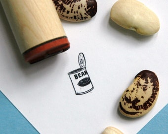 May include: A wooden stamp with a red rim next to a drawing of a bean can with the word "BEAN" on it. Several large beans with brown and tan markings are scattered around the stamp and drawing.