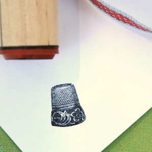 Op de afbeelding: Een houten stempel met een rode basis is boven een gestempelde afbeelding van een vingerhoed geplaatst. De afbeelding van de vingerhoed is zwart-wit, met een gedetailleerd bloemmotief rond de basis. Een lint met rode en witte strepen staat op de achtergrond.