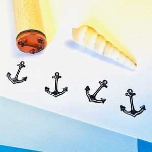 以下が含まれることがあります： アンカーデザインの木製スタンプと、4つのスタンプされたアンカー画像。貝殻も近くにあります。アンカーのスタンプは、クラフトやマリンをテーマにしたプロジェクトに適した、航海をテーマにしたものを作成します。