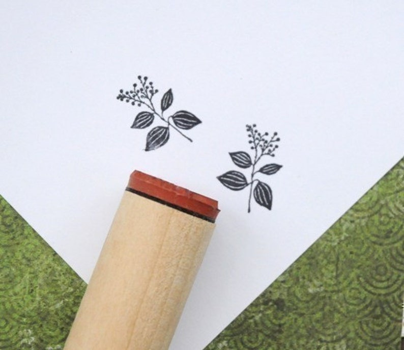 May include: A wooden stamp with a red rubber base and a botanical design. The stamp has a small sprig of leaves and berries. Two impressions of the stamp are visible on white paper. The background includes a green patterned paper.