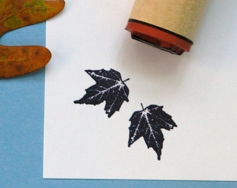 May include: A rubber stamp with a maple leaf design is shown next to two stamped black maple leaves on white paper. The stamp has a wooden handle and a red base. A dried leaf is in the corner on a blue background.