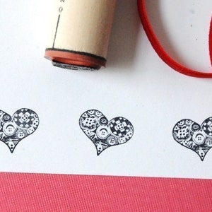 Może przedstawiać: Zbliżenie stempla gumowego i odbitych kształtów serca. Stempel wykonany jest z drewna i gumy. Kształty serca wypełnione są wzorami guzików. Czerwona wstążka znajduje się w tle. Serca są wydrukowane na białym papierze.