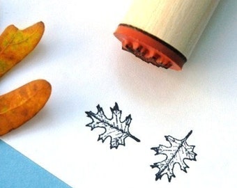 May include: A wooden handled stamp with a leaf design, next to two stamped leaf images. The stamp has an orange base. Two dried orange leaves are in the background. The stamped leaves are black.