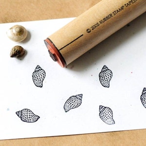 Puede incluir: Un sello de madera con un diseño de concha se muestra junto a varias impresiones estampadas del mismo diseño. También hay dos pequeñas conchas. El mango del sello tiene el texto "© 2005 RUBBER STAMP TAPESTRIES".