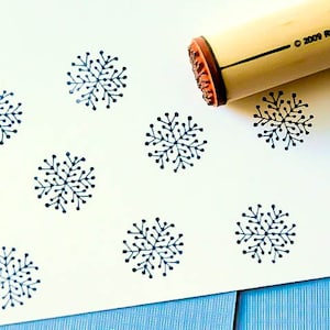 May include: A wooden rubber stamp with a snowflake design is shown next to several stamped impressions on white paper. The stamp handle is light brown, and the snowflake impressions are dark blue. The paper is on a light blue surface.