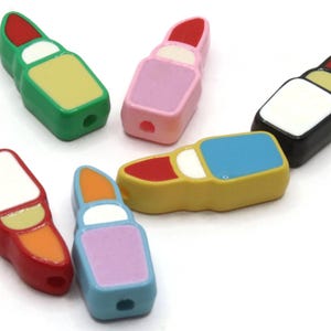 May include: A selection of lipstick-shaped beads in a variety of colours. The beads are red, green, pink, yellow, blue, and orange. Each bead has a white accent and a hole for threading, suitable for jewellery making.