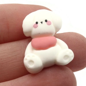 May include: White ceramic figurine of a dog wearing a pink collar. The dog has a cartoon-like face with black eyes and a pink nose.