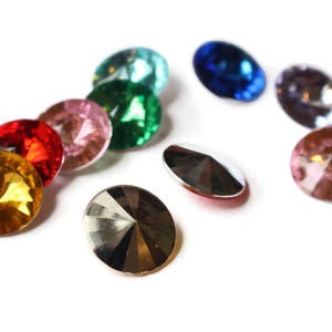 May include: Assortment of colorful glass rhinestones in various shapes and sizes. The rhinestones are arranged in a semi-circle on a white background.
