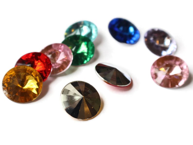 May include: Assortment of colorful glass rhinestones in various shapes and sizes. The rhinestones are arranged in a semi-circle on a white background.