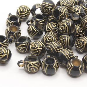 May include: A collection of black and gold decorative beads. Each bead is shaped like a small cup with a handle and features a gold swirl design. The beads are scattered on a white surface, creating a visually appealing arrangement.