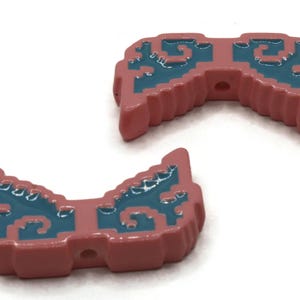 May include: Two pink, pixelated, decorative objects with teal accents. Each has a curved shape with a series of holes. The objects are on a white background.