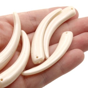 May include: Several off-white, curved pendants with a glossy finish. Each pendant has a small hole drilled at one end. The pendants are held in a hand, suggesting they are jewelry components or decorative elements.