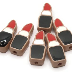 May include: Six lipstick-shaped beads. Each bead features a red lipstick tip, a white band, and a black base. The beads are a light brown color and have a hole for stringing. These beads are ideal for jewelry making.