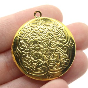 Op de afbeelding: Een goudkleurig rond medaillon met een bloemenontwerp. Het medaillon heeft een gedetailleerd reliëfpatroon van bloemen en bladeren. Het heeft een kleine lus aan de bovenkant om aan een ketting of koord te bevestigen. Het medaillon wordt in een hand gehouden.