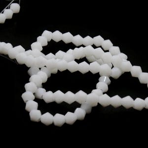 May include: Strands of white, faceted beads against a black background. The beads are strung together, forming multiple loops. The beads have a geometric, pentagonal shape. The image is well-lit, highlighting the beads' texture and color.