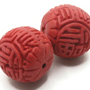 Op de afbeelding: Twee ronde rode kralen met ingewikkelde gegraveerde ontwerpen. De kralen zijn gemaakt van een glad, gepolijst materiaal en hebben een dieprode kleur.