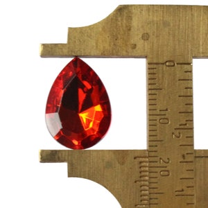 May include: A red teardrop-shaped crystal is measured with a brass caliper. The caliper is marked in inches.