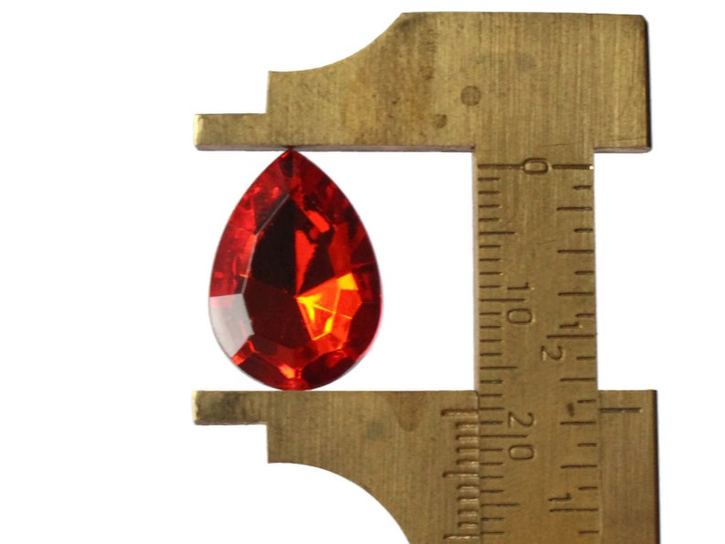 May include: A red teardrop-shaped crystal is measured with a brass caliper. The caliper is marked in inches.