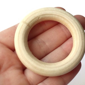 May include: A natural wood ring, approximately 2 inches in diameter, with a smooth, rounded surface. The ring is shown in a hand, demonstrating its size and shape.