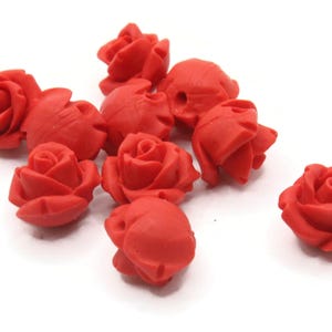Op de afbeelding: Een verzameling rode roosvormige kralen. De kralen zijn verspreid over een witte ondergrond. De rozen bevinden zich in verschillende bloeistadia, met gedetailleerde bloembladformaties.