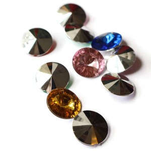 May include: Assortment of colorful, faceted, round glass rhinestones with silver backing. The rhinestones are arranged in a cluster on a white background.
