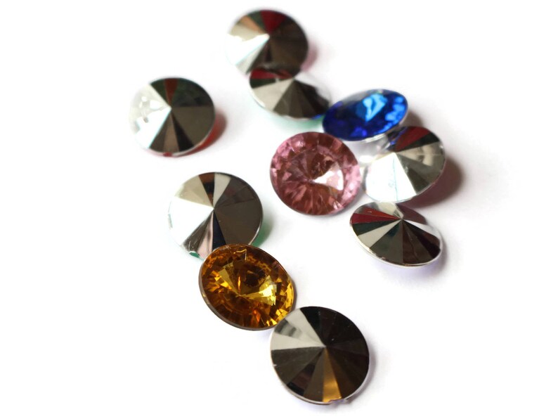 May include: Assortment of colorful, faceted, round glass rhinestones with silver backing. The rhinestones are arranged in a cluster on a white background.