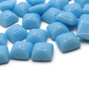 May include: A group of light blue glass square beads with a slightly rounded edge. The beads are scattered on a white surface.