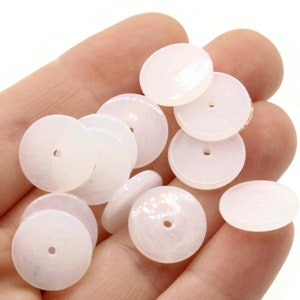 May include: A collection of white, round, flat, shell beads with a hole in the centre. The beads are approximately 2.5 cm in diameter.