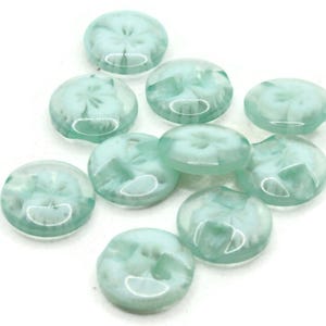 Puede incluir: Una colección de diez botones redondos, translúcidos, de color verde claro con un diseño de remolino blanco.