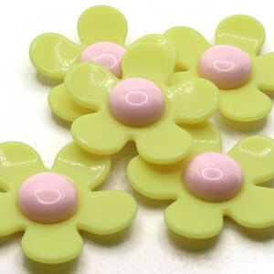 May include: A collection of yellow daisy-shaped embellishments with pink centers. These plastic flower accents have a glossy finish and are arranged on a white surface. Ideal for craft projects, scrapbooking, or adding a floral touch.