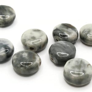 Op de afbeelding: Een verzameling van acht ronde, platte, keramische kralen met een wit en grijs marmerpatroon. De kralen hebben een gat dat door het midden is geboord om te rijgen.