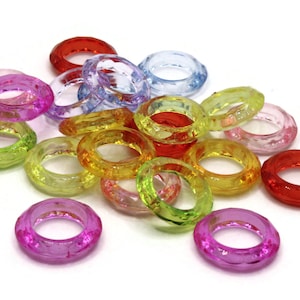 May include: A collection of colorful, faceted acrylic rings in various shades including red, pink, green, yellow, and blue. The rings are transparent and appear to be decorative or for crafting purposes. The rings are scattered on a white surface.