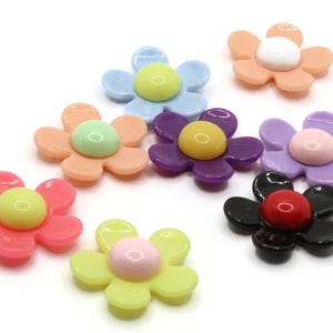 Puede incluir: Una colección de adornos en forma de flor colorida. Las flores presentan varios colores, incluyendo rosa, amarillo, azul, morado y negro, con acentos centrales contrastantes. Estos elementos decorativos son adecuados para manualidades y adornos.