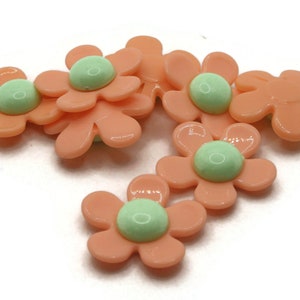 May include: Several peach-coloured flower-shaped embellishments with light green centres. The petals are smooth and glossy, and the centres have a rounded, button-like appearance. These decorative elements are suitable for craft projects.