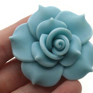 May include: A light blue fabric flower with a rosette center. The flower has five petals and is made of a soft, pliable material.