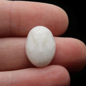 Puede incluir: Pequeña talla ovalada de piedra blanca que representa el rostro de un faraón. La talla está detallada con rasgos faciales y un tocado. La piedra se sostiene entre los dedos sobre un fondo negro.