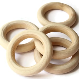 May include: Five unfinished wooden rings, each about 7.6 cm in diameter, are displayed. The rings are a light beige hue, suggesting natural wood with visible grain. They are arranged overlapping against a white backdrop, ideal for craft projects or toys.