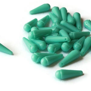 May include: A group of teal-colored glass teardrop beads. The beads are all the same size and shape, and they are arranged in a pile on a white surface.