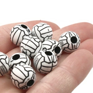 May include: A close-up of a hand holding a group of white plastic beads with black geometric designs. The beads have a hole in the center for stringing.