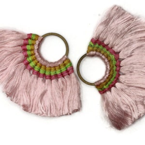 May include: A pair of pink tassel earrings with a woven design in green, pink, and gold. The earrings have a circular metal hoop.