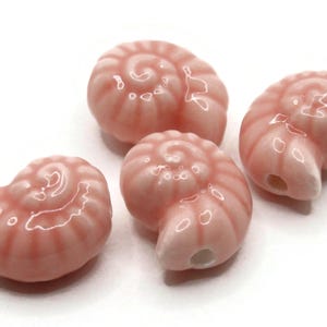 Puede incluir: Cuatro cuentas de cerámica rosa en forma de concha con un acabado brillante. Las cuentas tienen forma de concha con un patrón en espiral.