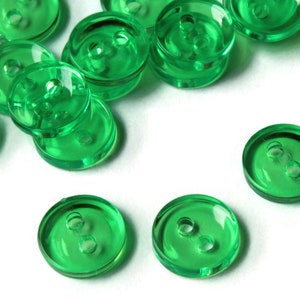 Puede incluir: Un conjunto de botones redondos verdes transparentes, cada uno con dos agujeros. Los botones están hechos de un material brillante y están dispersos sobre una superficie blanca. Los botones son de un tono verde vibrante.