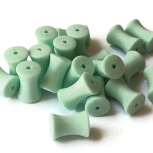 May include: A group of 17 light green, ceramic beads with a cylindrical shape and a hole in the center. The beads are arranged in a pile on a white surface.