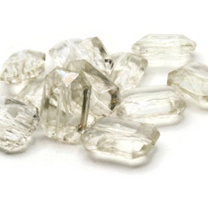May include: A group of clear glass beads with a faceted, rectangular shape. The beads are scattered on a white surface.
