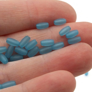 May include: A handful of light blue, oval-shaped beads. The beads have a frosted, matte finish and are scattered across the palm of a hand. The beads are small, likely for jewelry making or crafts.