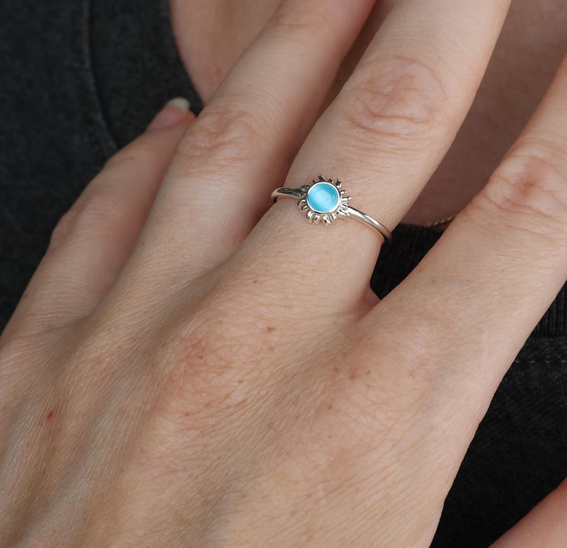 May include: A silver ring with a blue gemstone in the center, surrounded by a sunburst design.